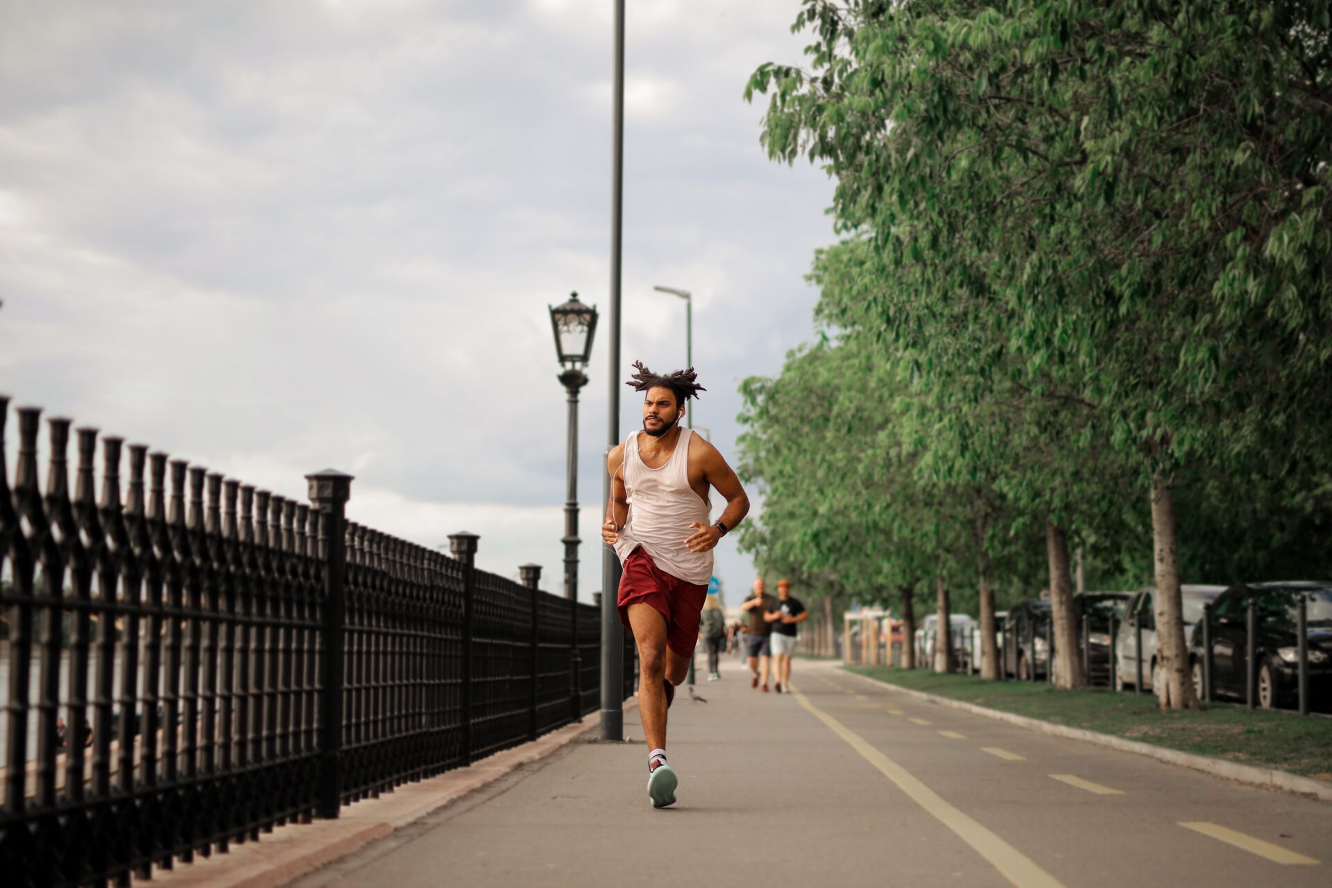 Runner in a white tank top and red shorts mid-stride on a fenced waterfront path, mouth open, earbud cable visible on his chest.