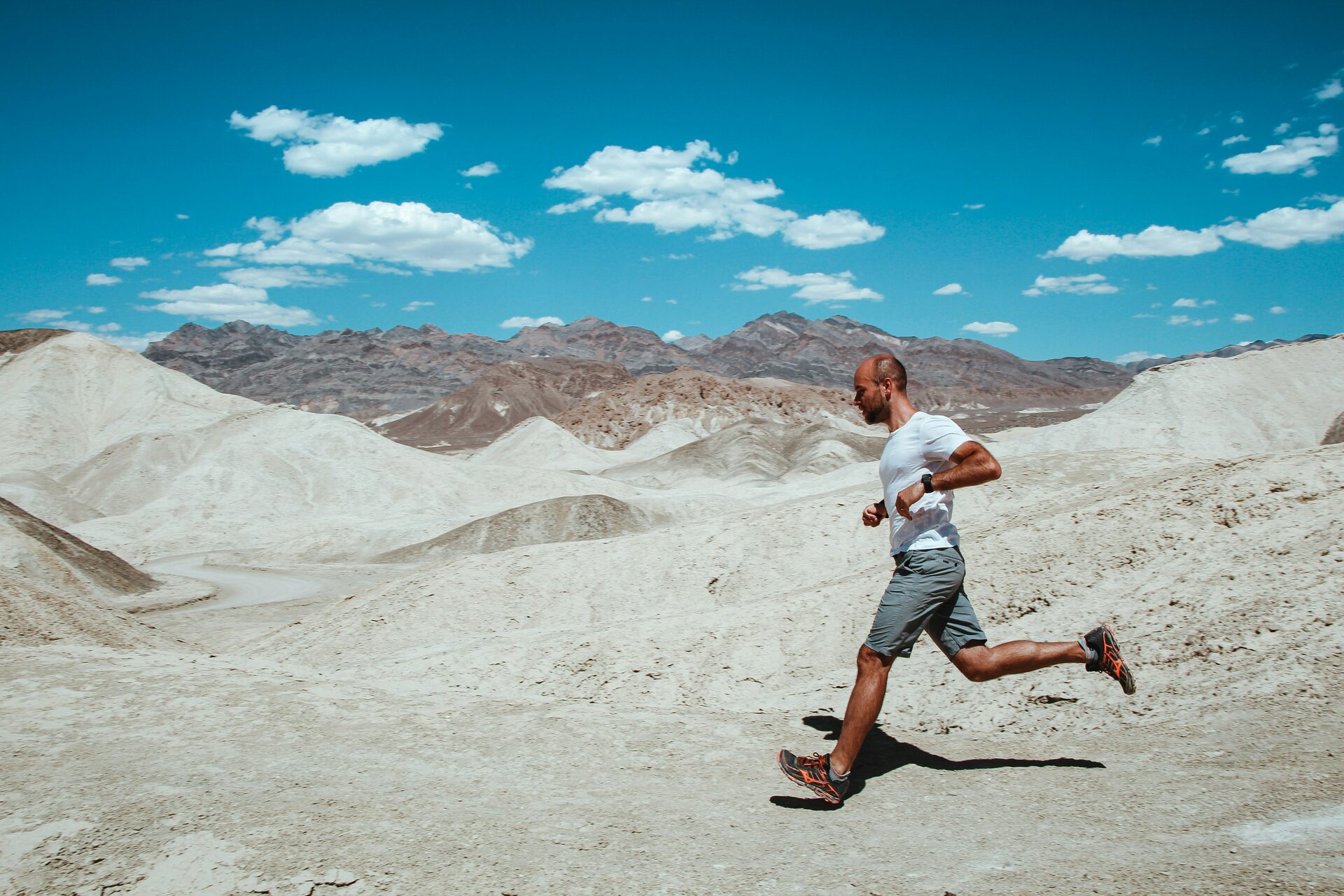 A runner in a white t-shirt and gray shorts mid-stride across pale desert hills under a bright midday sky, bare mountains behind.