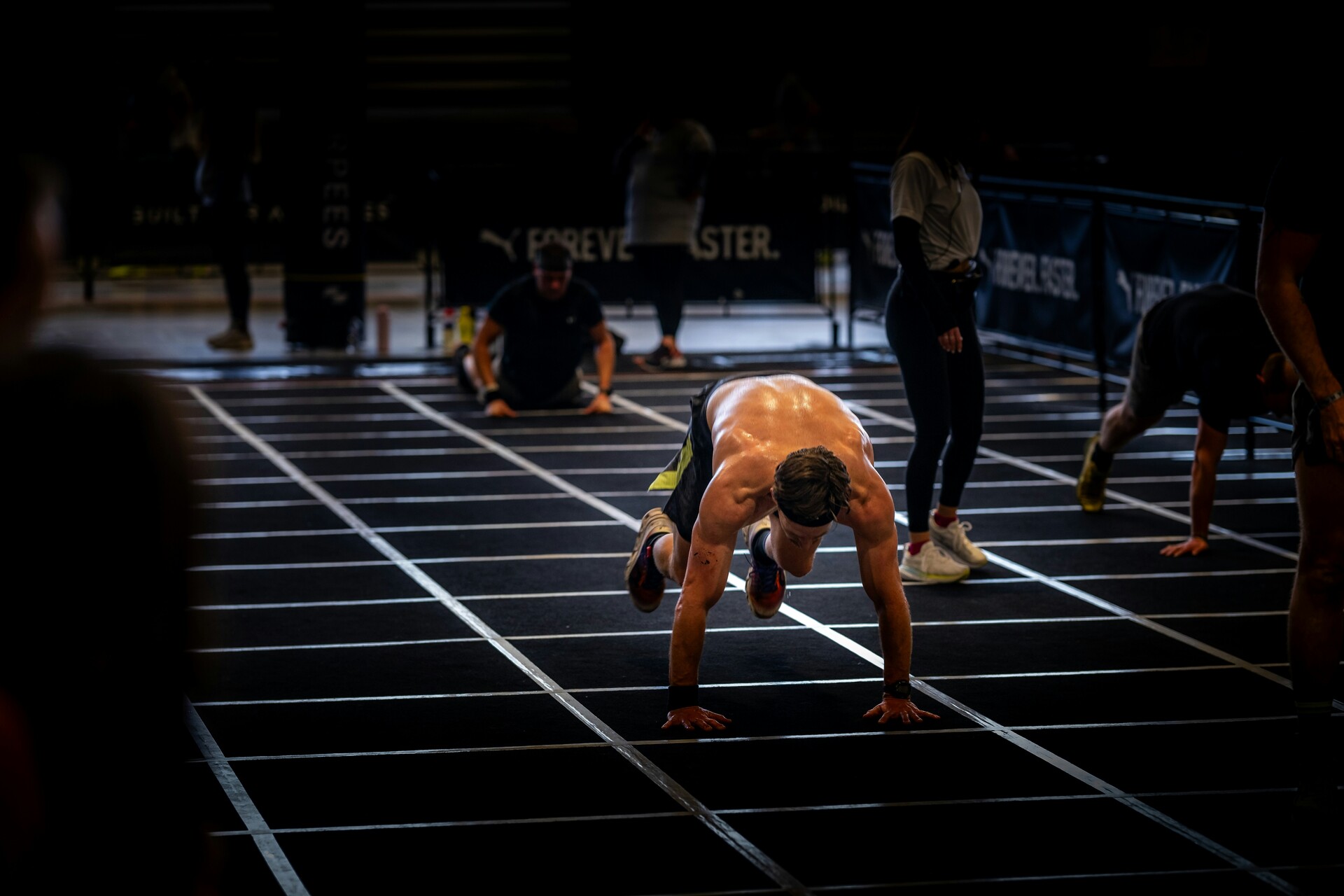 An athlete mid burpee broad jump on a taped indoor race floor, hands down on the lane, with other athletes working their way down the lanes behind.