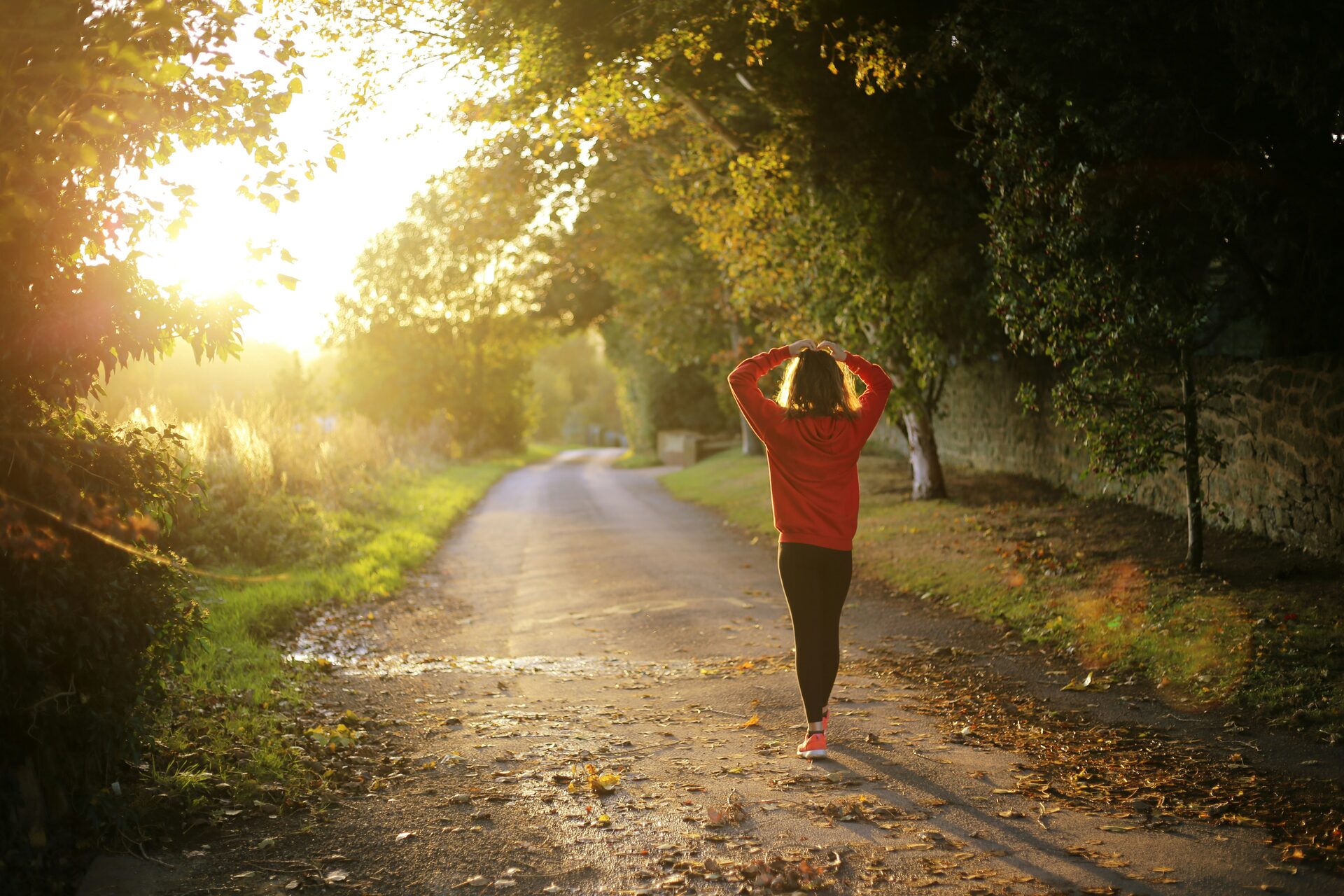 A runner walking along a tree-lined lane in low sun, hands on her head.
