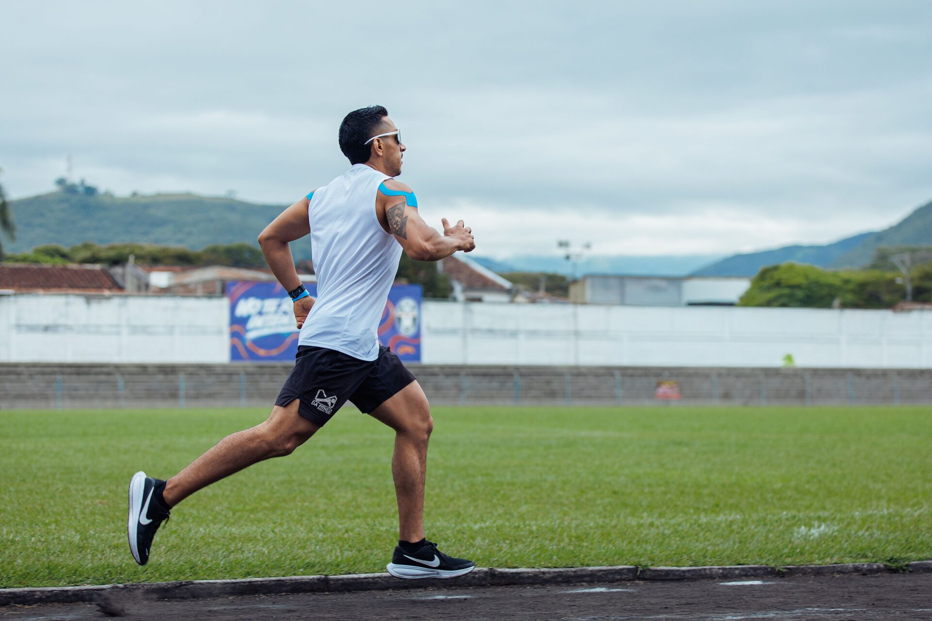 A runner in a white vest mid-stride down the straight of an outdoor track, alone, with a grass infield and cloud-covered hills behind.