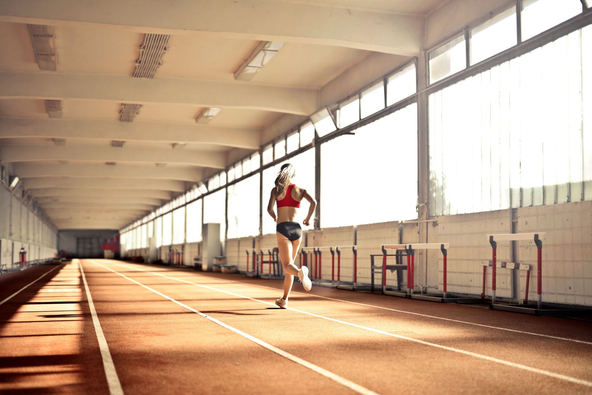 A runner in a red top running away down the lane lines of a sunlit indoor track, hurdles stacked along the wall beside her.