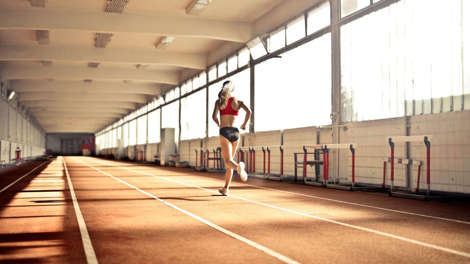 A runner in a red top running away down the lane lines of a sunlit indoor track, hurdles stacked along the wall beside her.