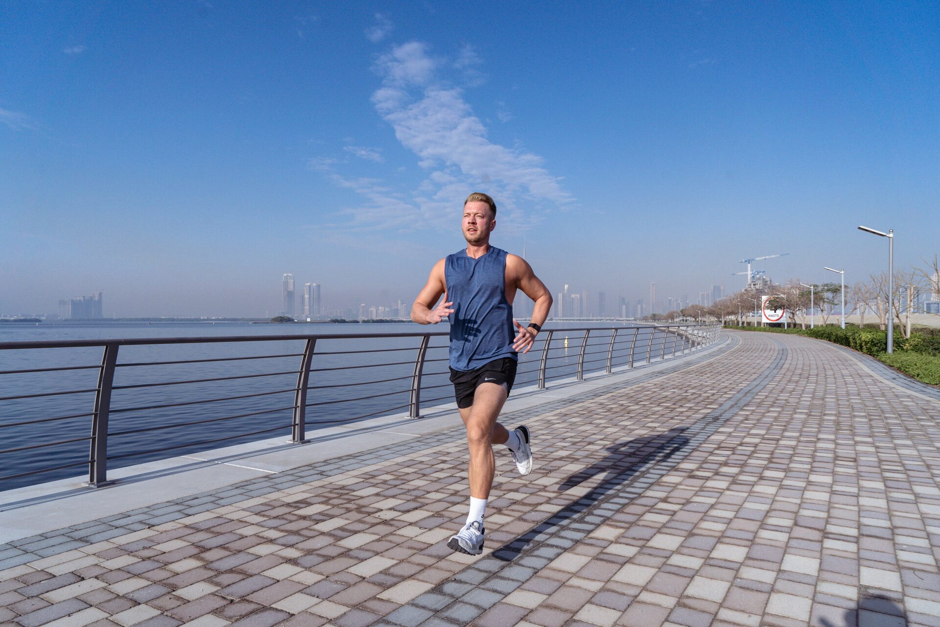 Runner in a sleeveless shirt mid-stride on a waterfront path, breathing hard, watch on his wrist.