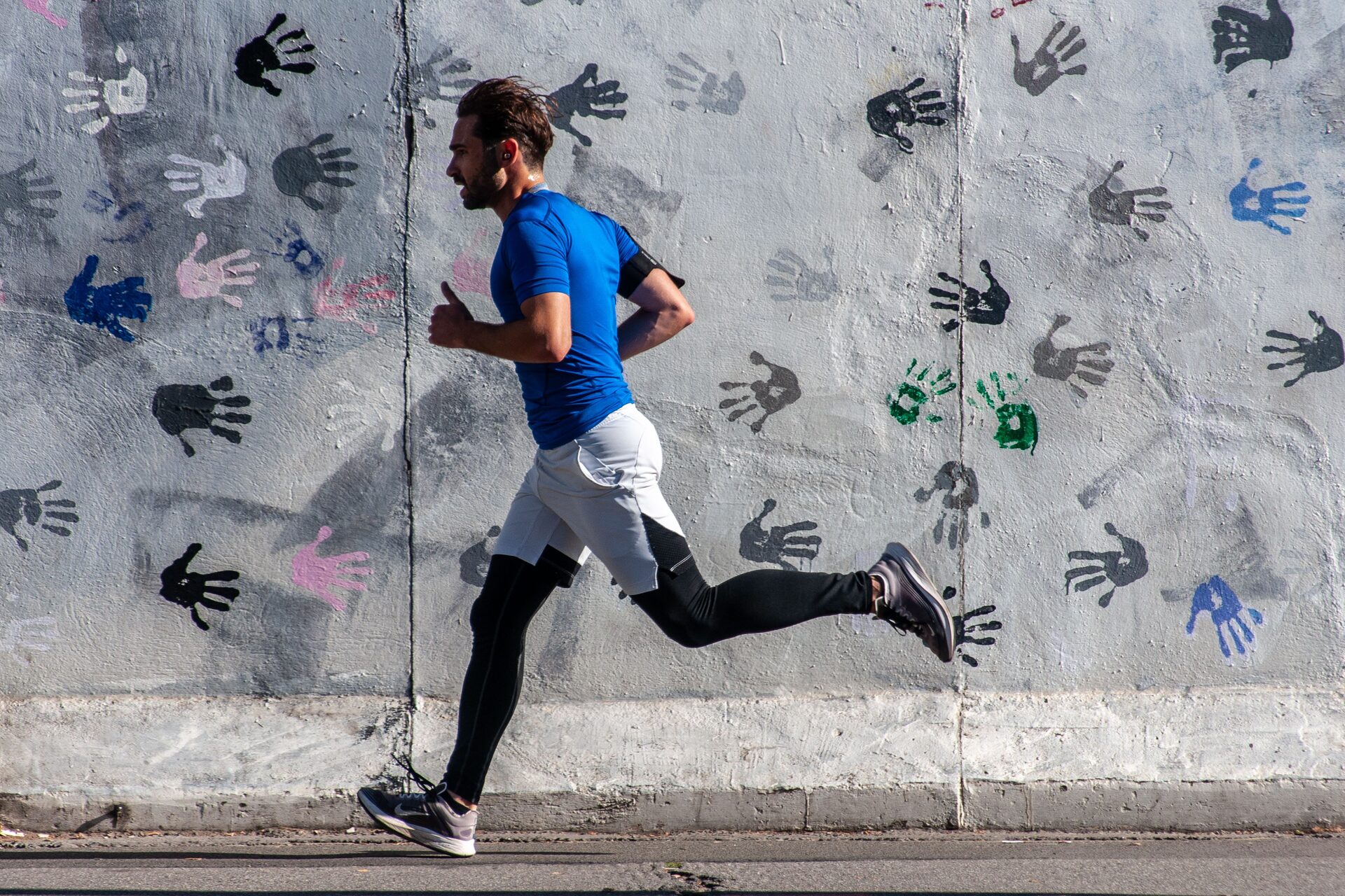 A runner mid-stride past a painted wall, working hard, earbud in, shot from the side.