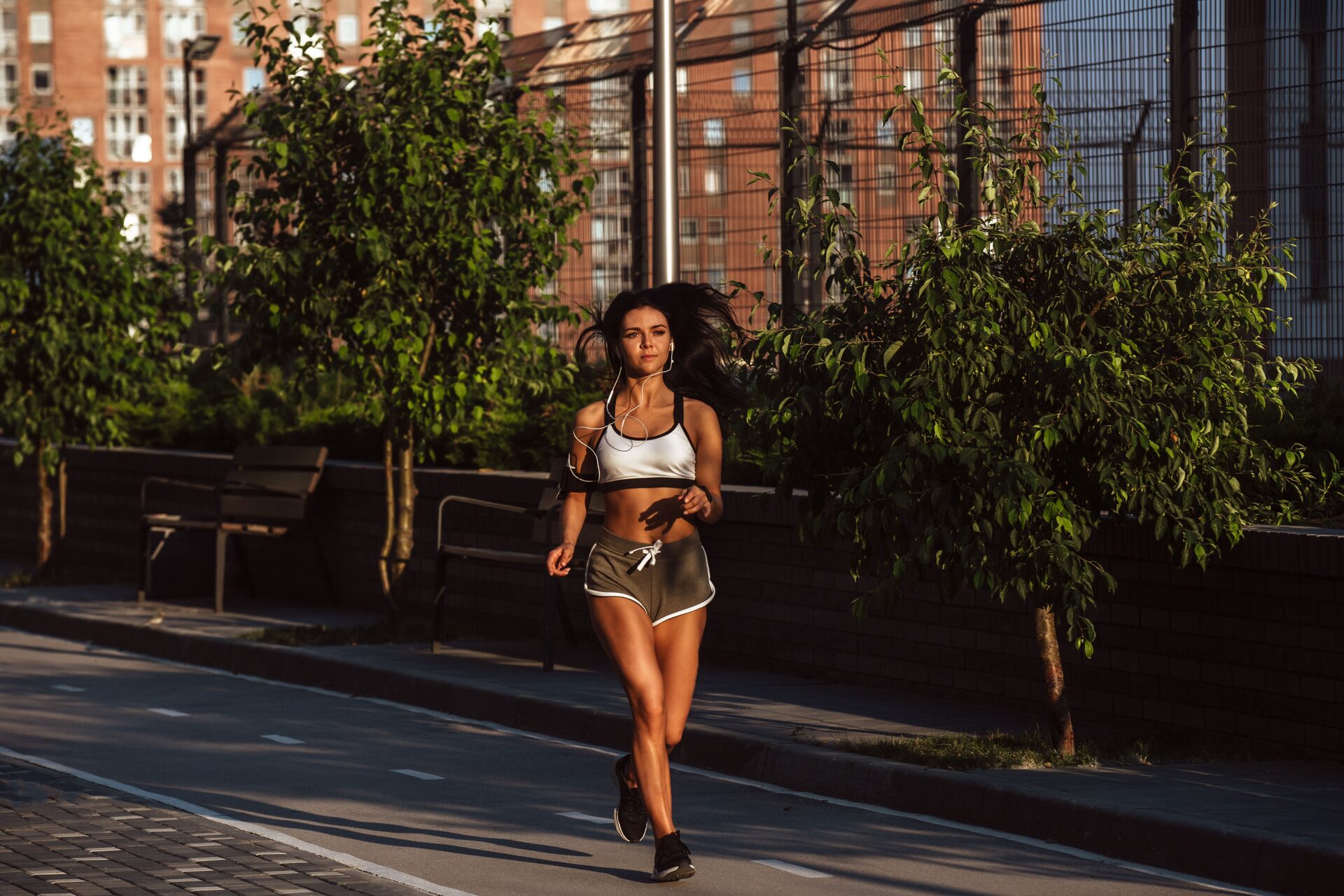 Runner in a white sports top and olive shorts on a city path in low evening sun, earphones in, hair lifted behind her, trees and a wire fence behind.