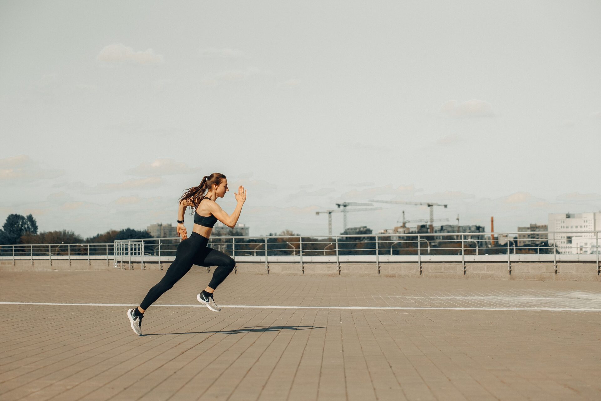Woman in a black sports bra and leggings sprinting on a paved rooftop running track, arms driving, both feet off the ground, construction cranes and city buildings in the background.