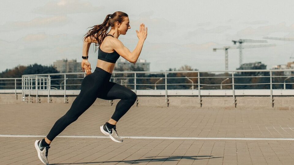 Woman in a black sports bra and leggings sprinting on a paved rooftop running track, arms driving, both feet off the ground, construction cranes and city buildings in the background.