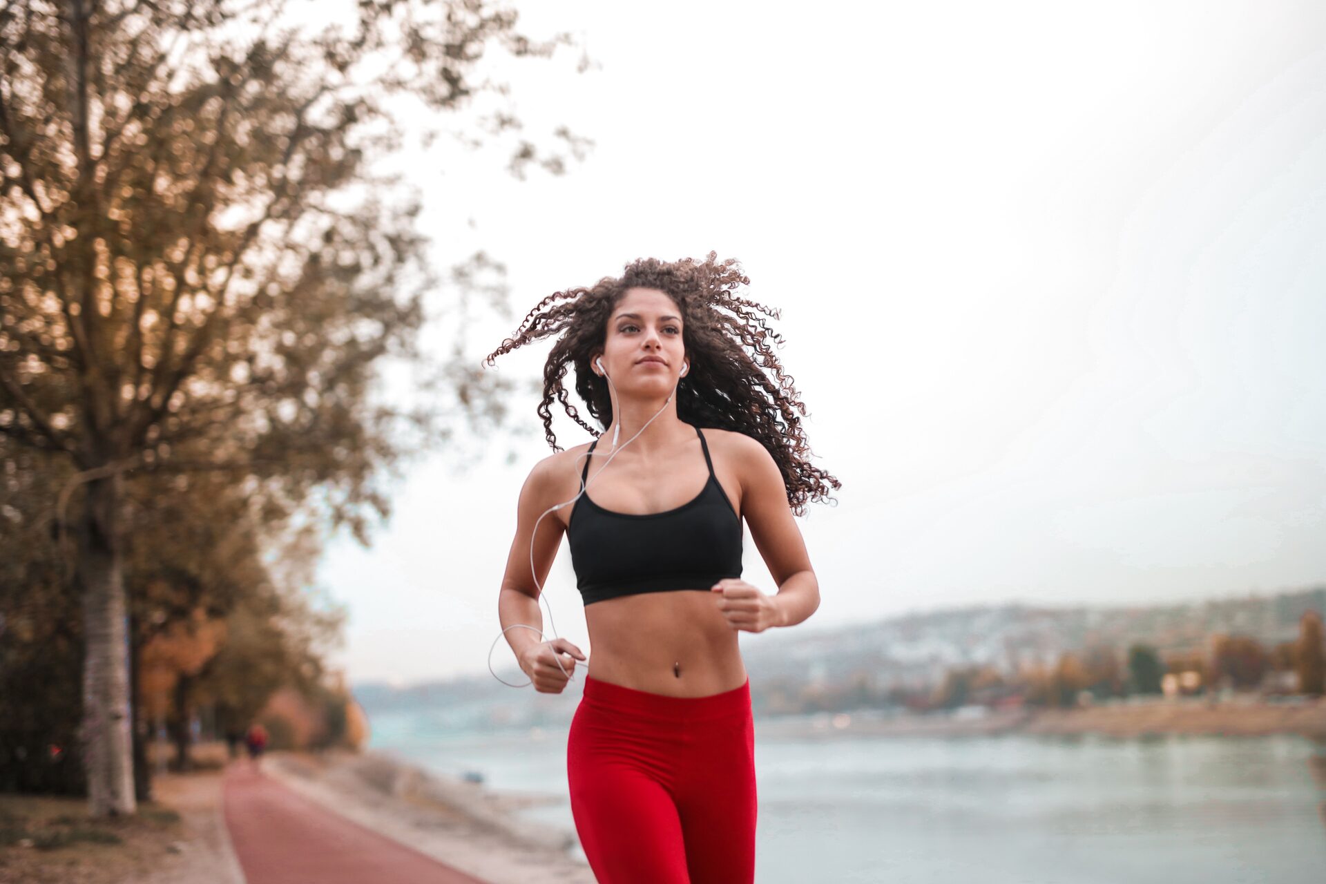 A runner in a black top and red leggings on a riverside path under an overcast sky, wired earphones in.