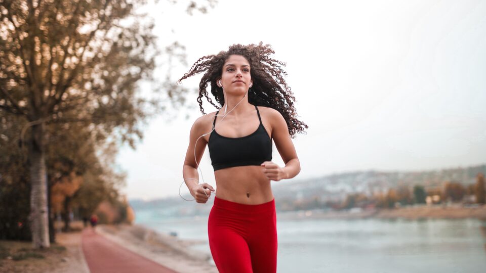 A runner in a black top and red leggings on a riverside path under an overcast sky, wired earphones in.