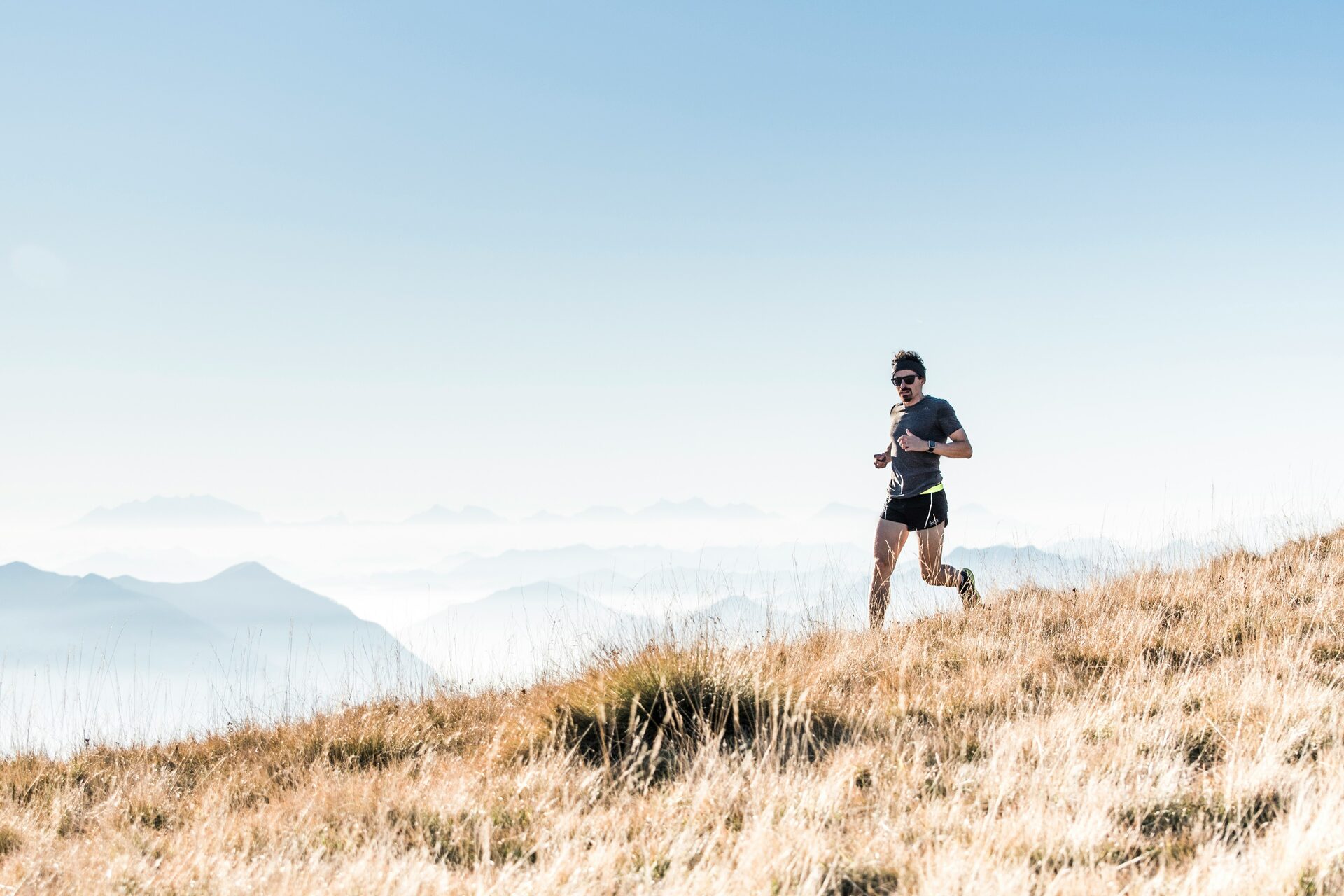 A runner in a gray t-shirt, sunglasses, and a dark headband crossing a slope of dry golden grass high above a sea of clouds, faint mountain ridgelines behind.