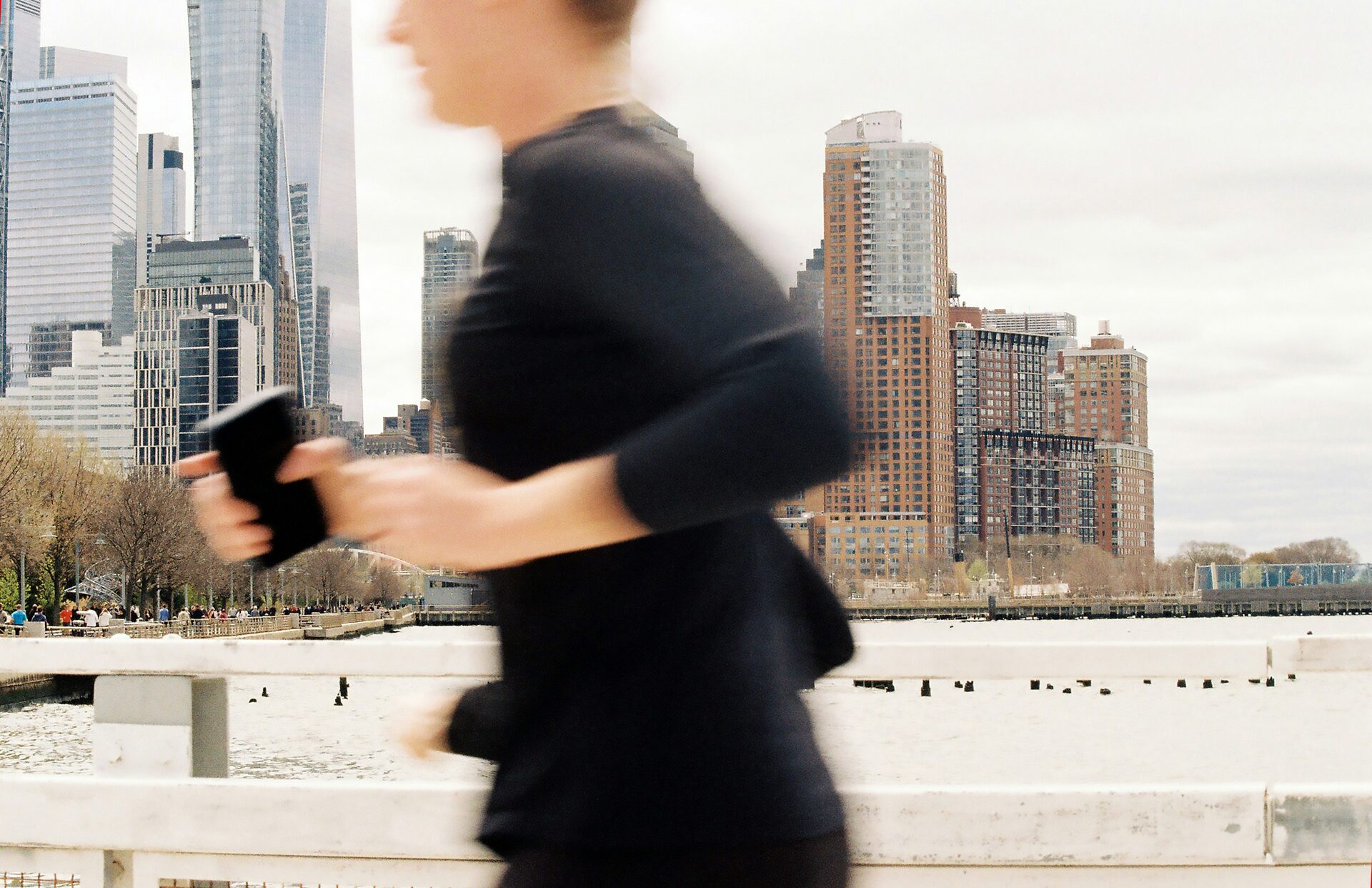 Runner passing a waterfront railing with a phone in hand, city skyline behind.