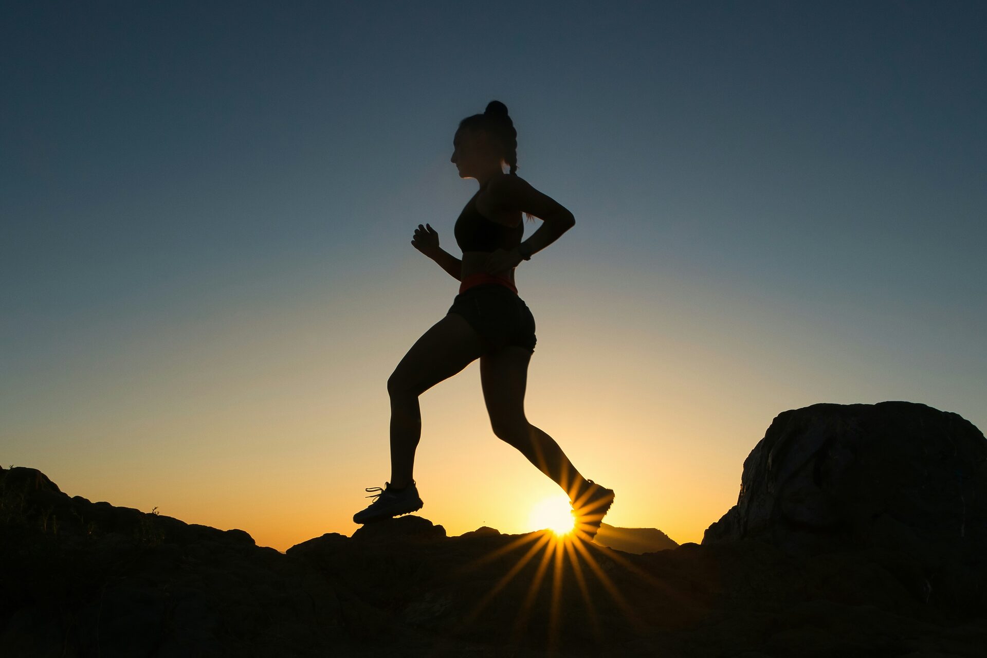 A runner in silhouette mid-stride along a rocky ridge at sunset, the sun sitting low between the runner's feet.